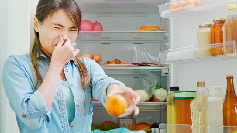 Woman holding her nose and an orange in front of open fridge