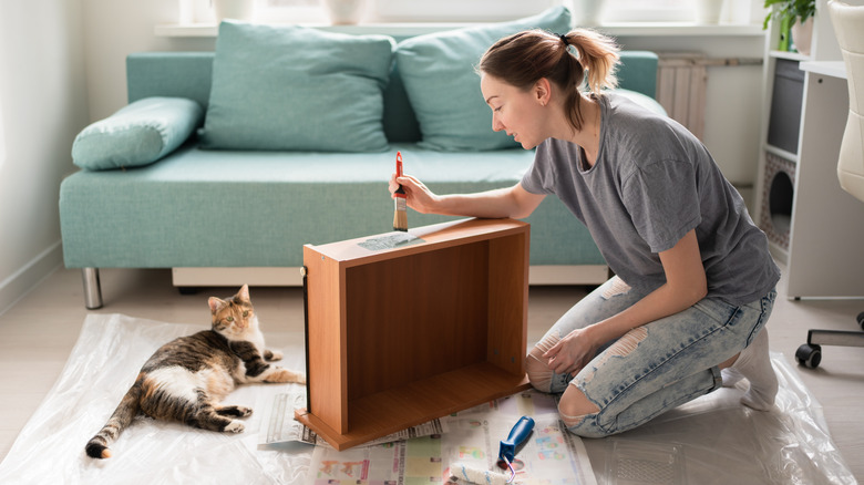 A woman with a short, brown ponytail paints a drawer set on newspaper in a living room.