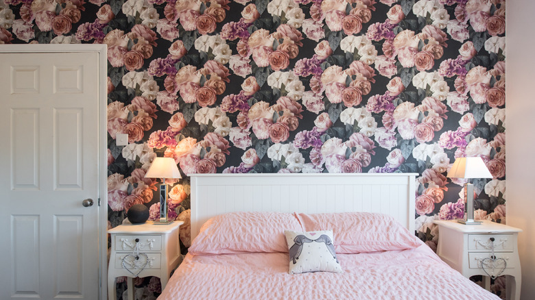 A bold floral wallpaper is hung on a wall behind a pink bed and white side tables