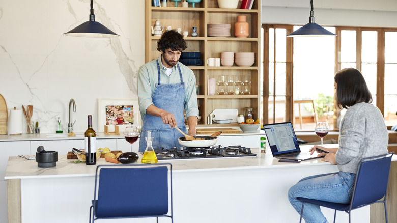 A man cooks at the kitchen island while a woman is on her laptop, seated at the island.