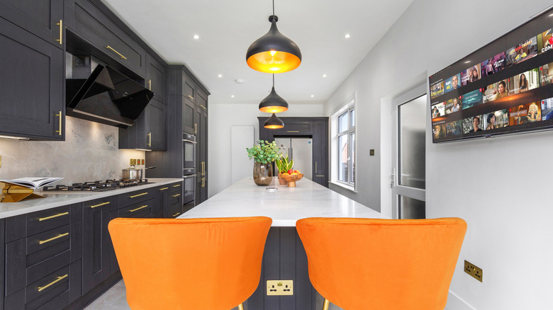 Two orange chairs sit at the end of a long white kitchen island, with three black and gold pendant lights overhead.