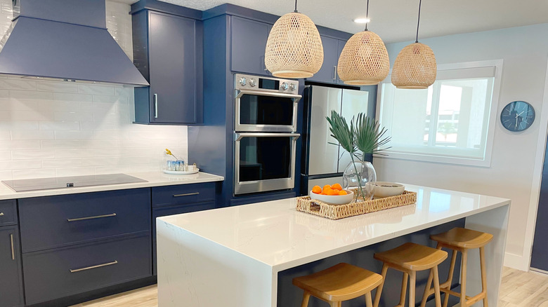 A minimalist kitchen features a white island topped with a wicker tray containing two shallow white bowls (one holding oranges) and a clear vase with two palm branches.