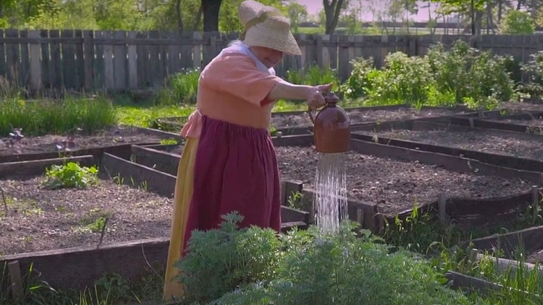 A reenactor uses a watering bell in a garden.