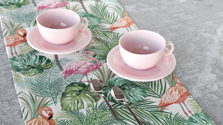 Two pink teacups sit on a tropical flamingo pattern Jacquard table runner