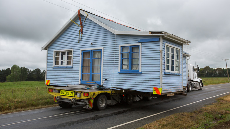 A blue wooden house is moved down the road on the back of a flatbed