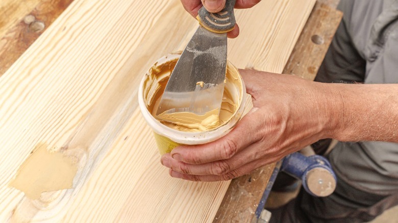 Man dipping putty knife in container of wood filler over wood board