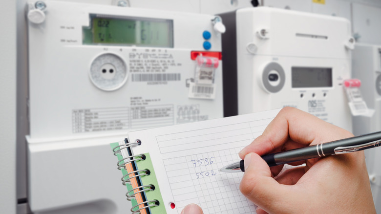 A close-up of a hand writing numbers in front of an electric meter