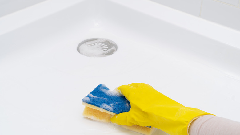 A person with a yellow rubber glove cleaning the bottom of an acrylic shower with a sponge