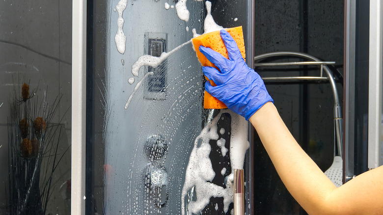 A gloved hand wiping glass shower door with sponge