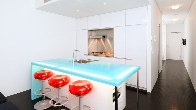 A modern kitchenette is fitted with a waterfall glass countertop illuminated by LED lights and three red stools beside the counter