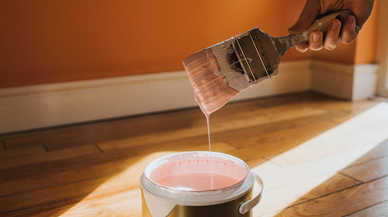 A hand holding a brush over a can of paint on a wood floor