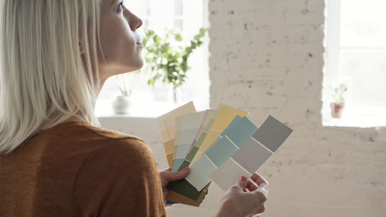 A woman looks a white brick walls while holding several paint color cards