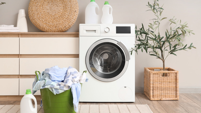 Laundry room with washing machine, a bucket with dirty clothes, detergents and a plant