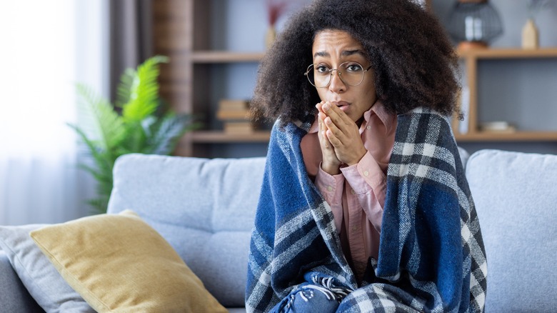 woman on couch wrapped in blanket breaths on hands for warmth