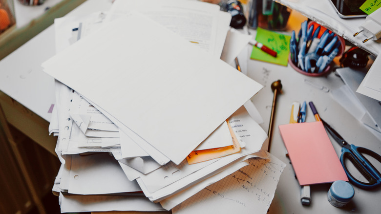 A desk space is cluttered with papers, pens, and other supplies