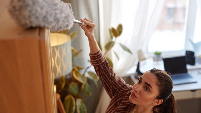 a woman dusting the top of a wooden cabinet